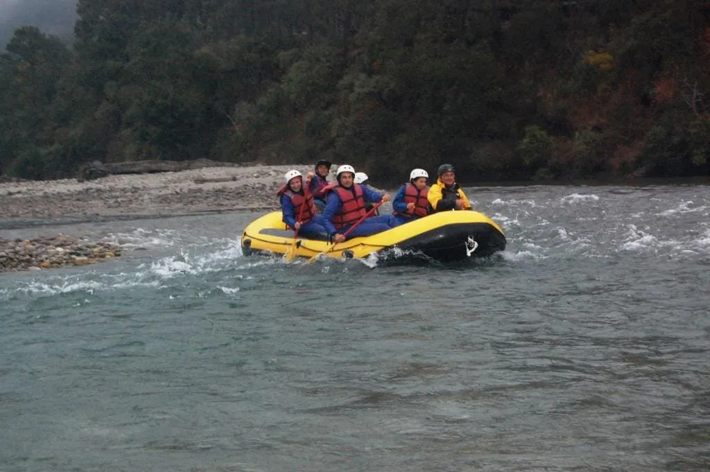 Six rafters in helmets and life jackets paddling a yellow raft through white water, with a stony bank and forested hillside behind