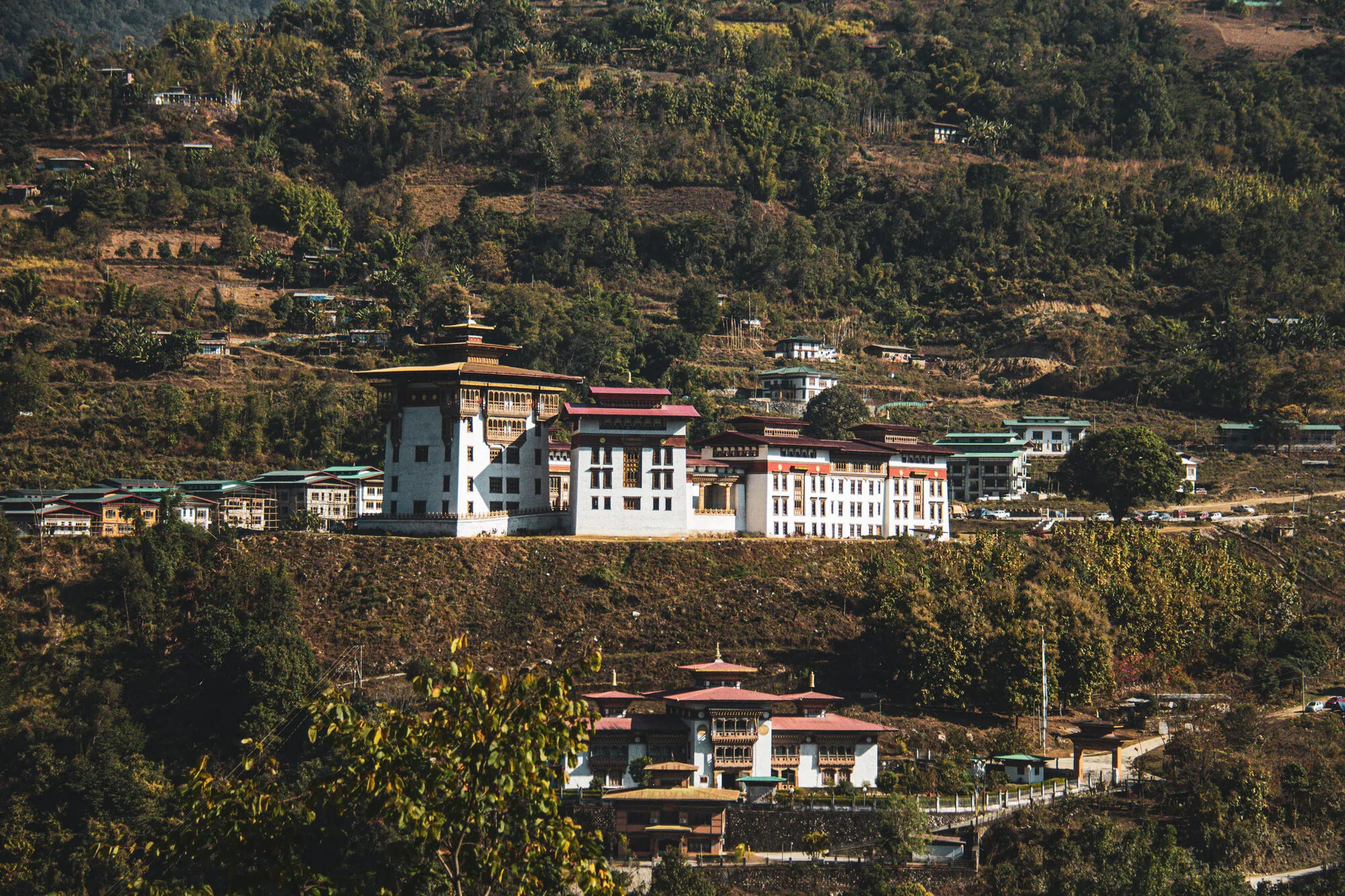 A walled building on a terrace at the lip of a steep bank, a second walled complex with a tower below it, and terraced slopes of houses and new blocks behind, seen across the valley