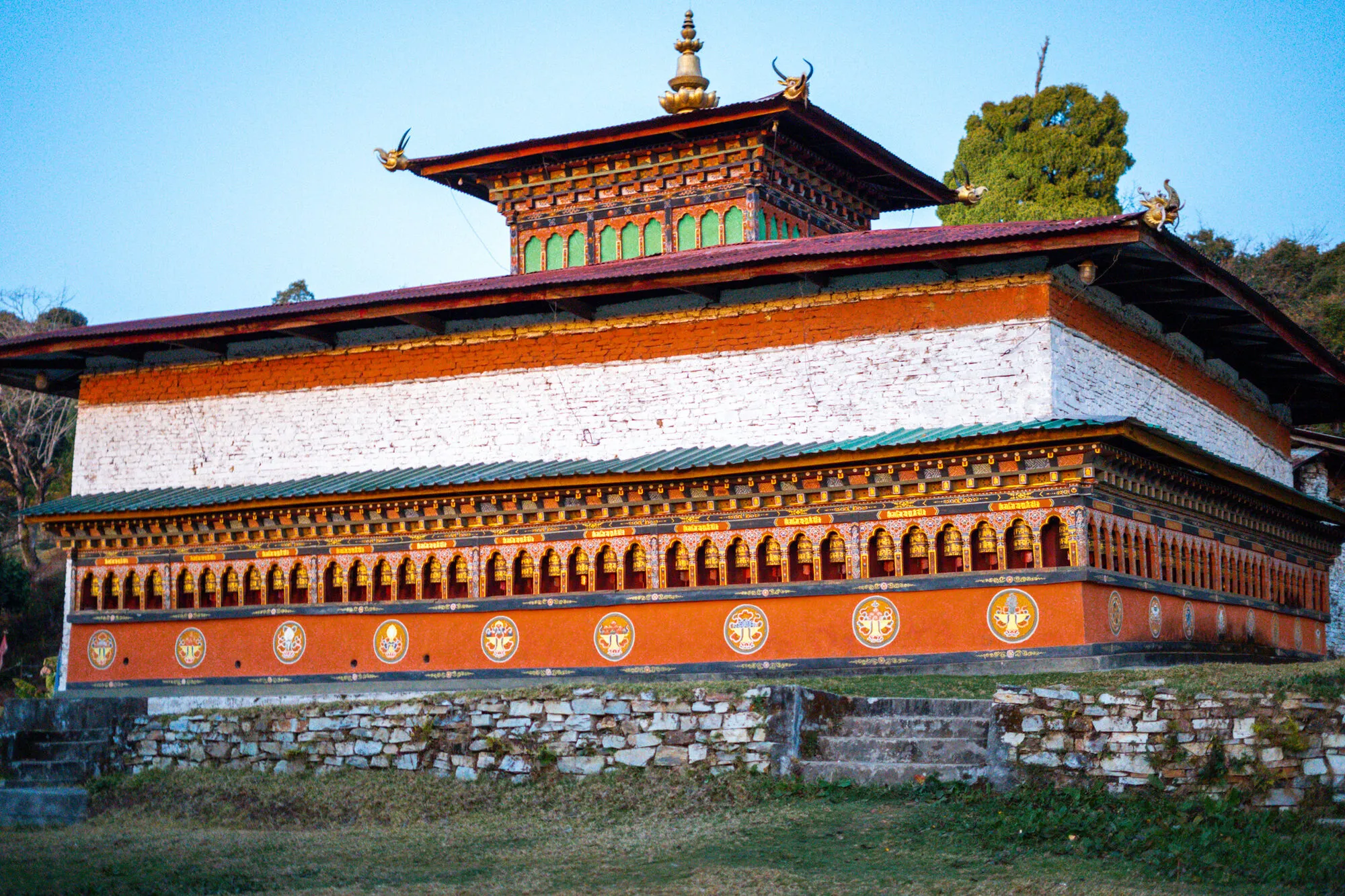 A long temple at Dungkhar Goenpa with a gallery of prayer wheels along its orange lower storey, a red roof and a green-windowed lantern storey, cypresses behind