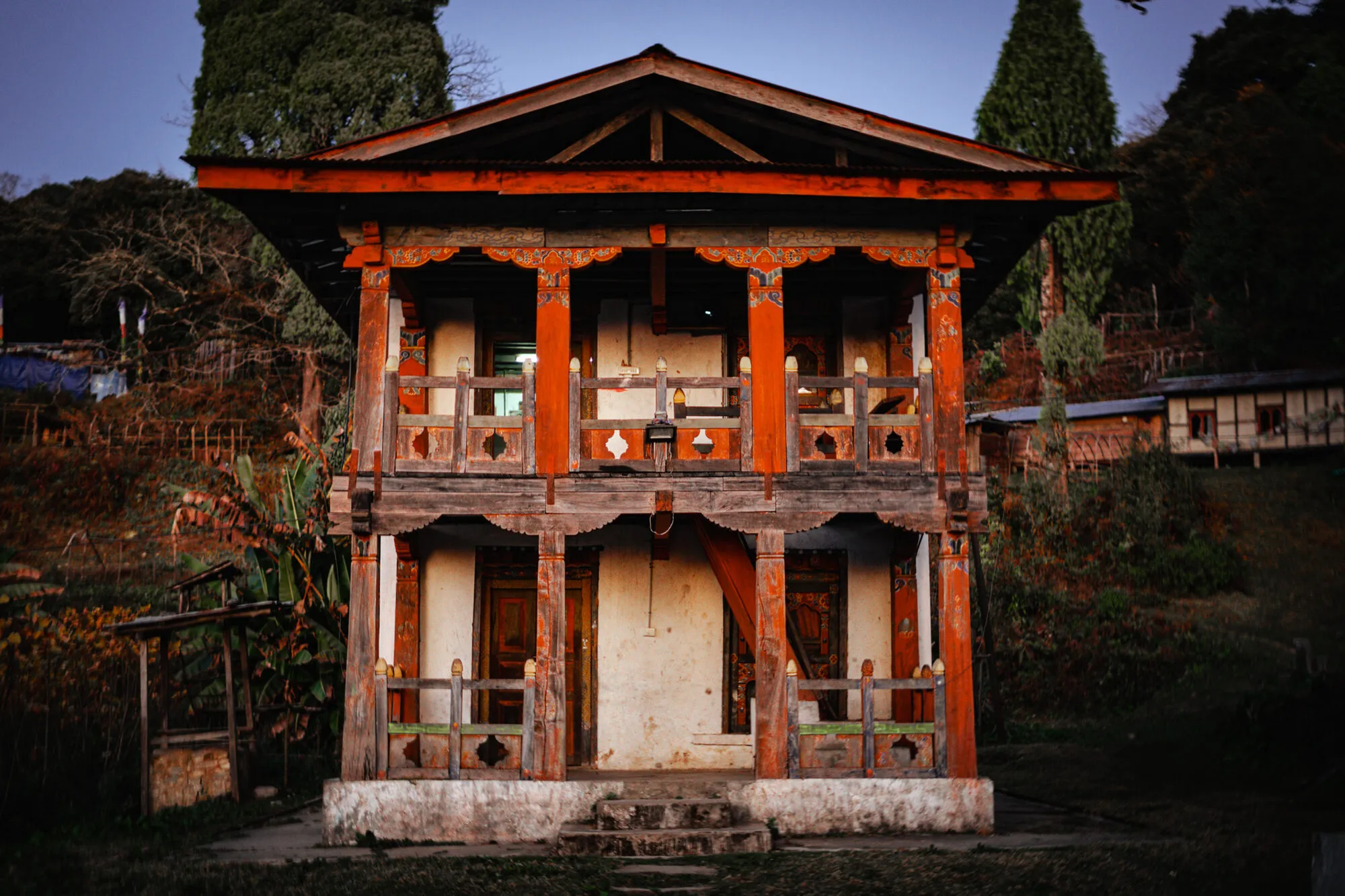 A two-storey wooden-fronted building with red pillars and railings at Dungkhar Goenpa at dusk, cypress trees behind and village houses on the slope