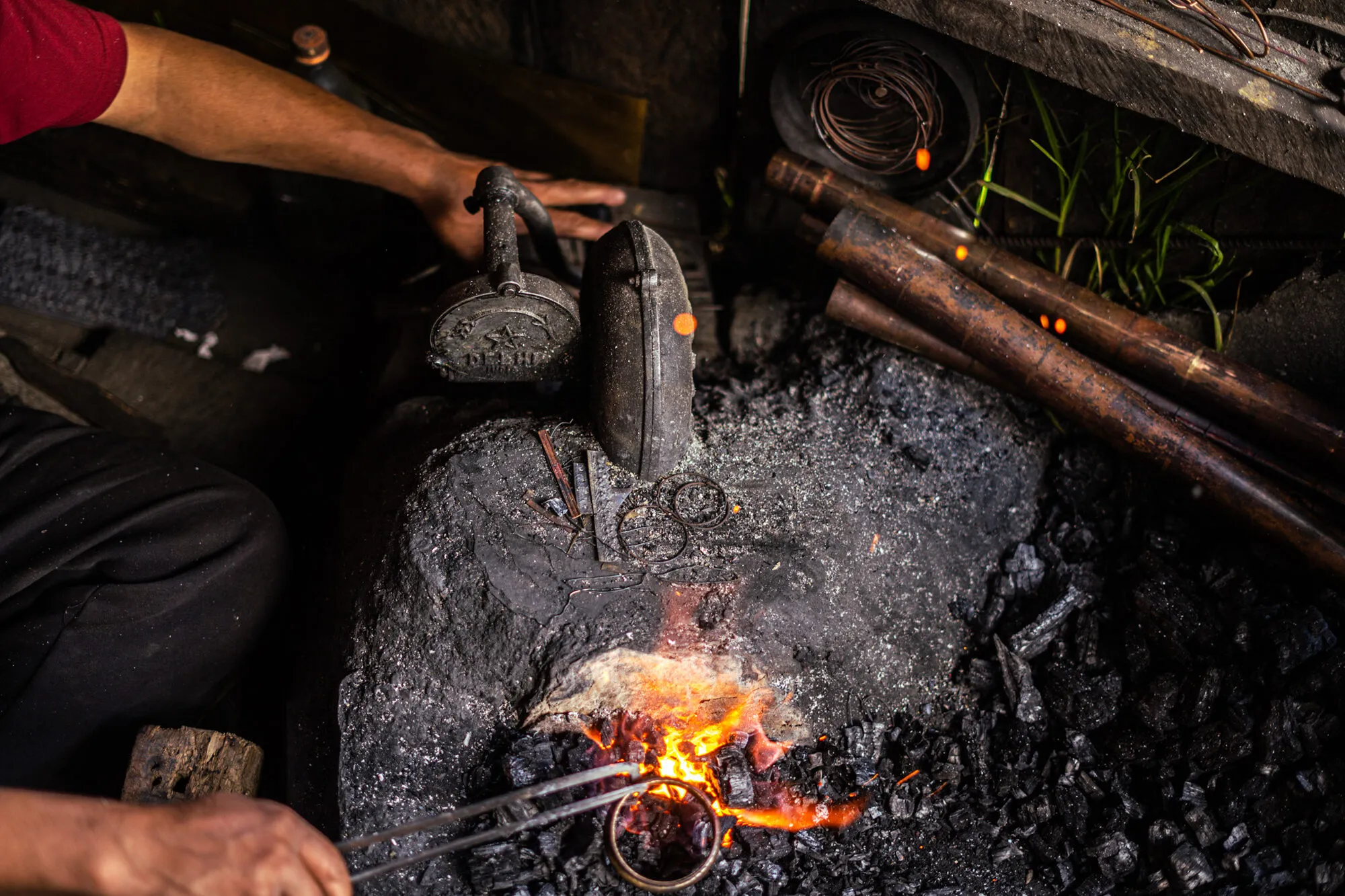 A charcoal forge from above: a metal ring held in tongs over glowing coals, copper tube stock in the fire and a coil of copper wire beside it