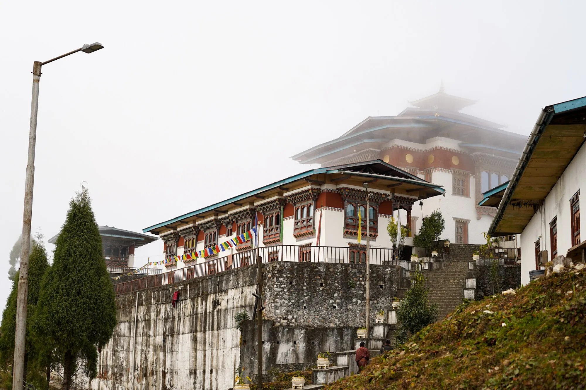 The main lhakhang at Yongla Goenpa in mist above its high stone retaining wall, with prayer flags and stone steps in front