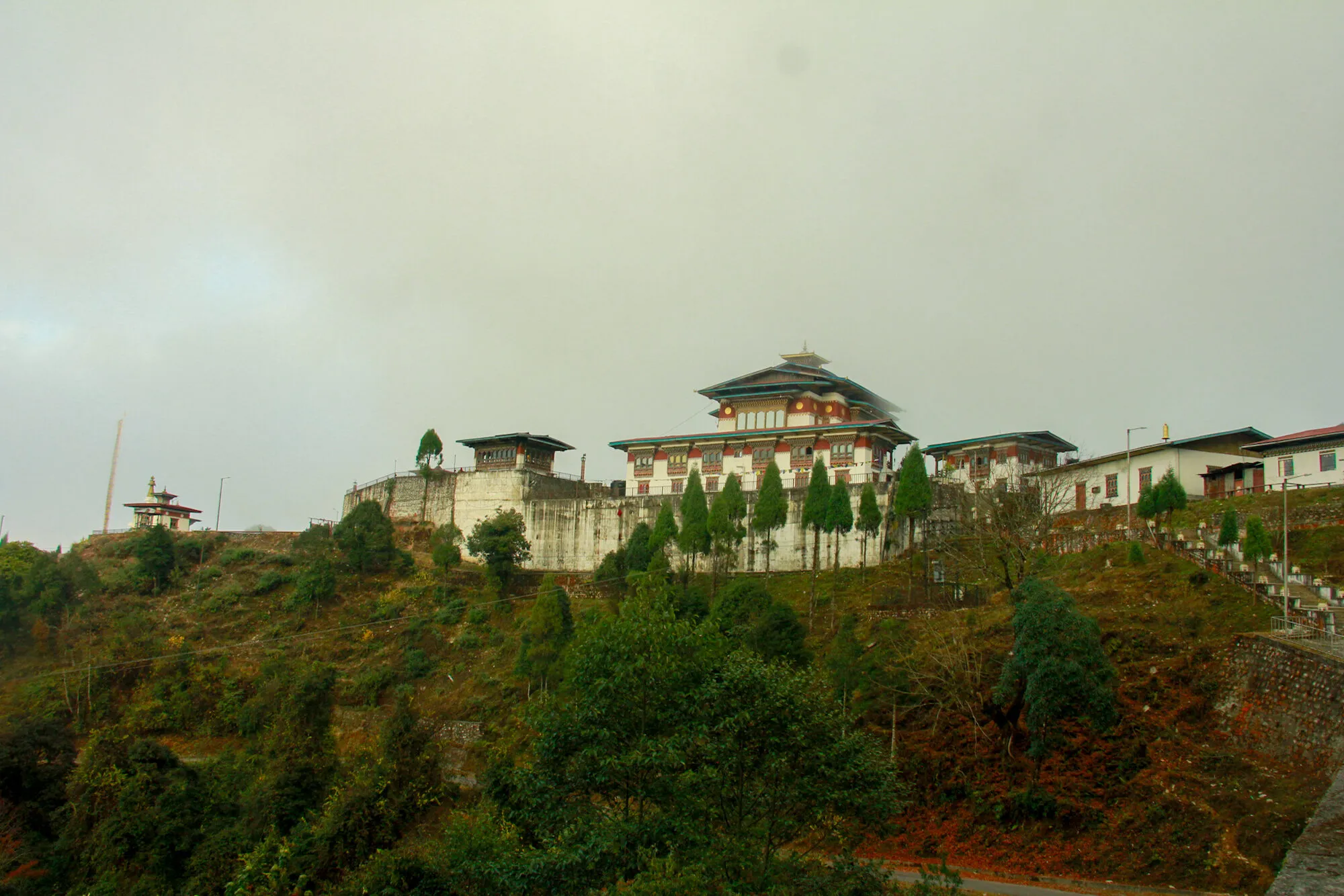 Yongla Goenpa on its ridge seen from the road below: the retaining wall, the lhakhang, a row of cypresses and the side buildings