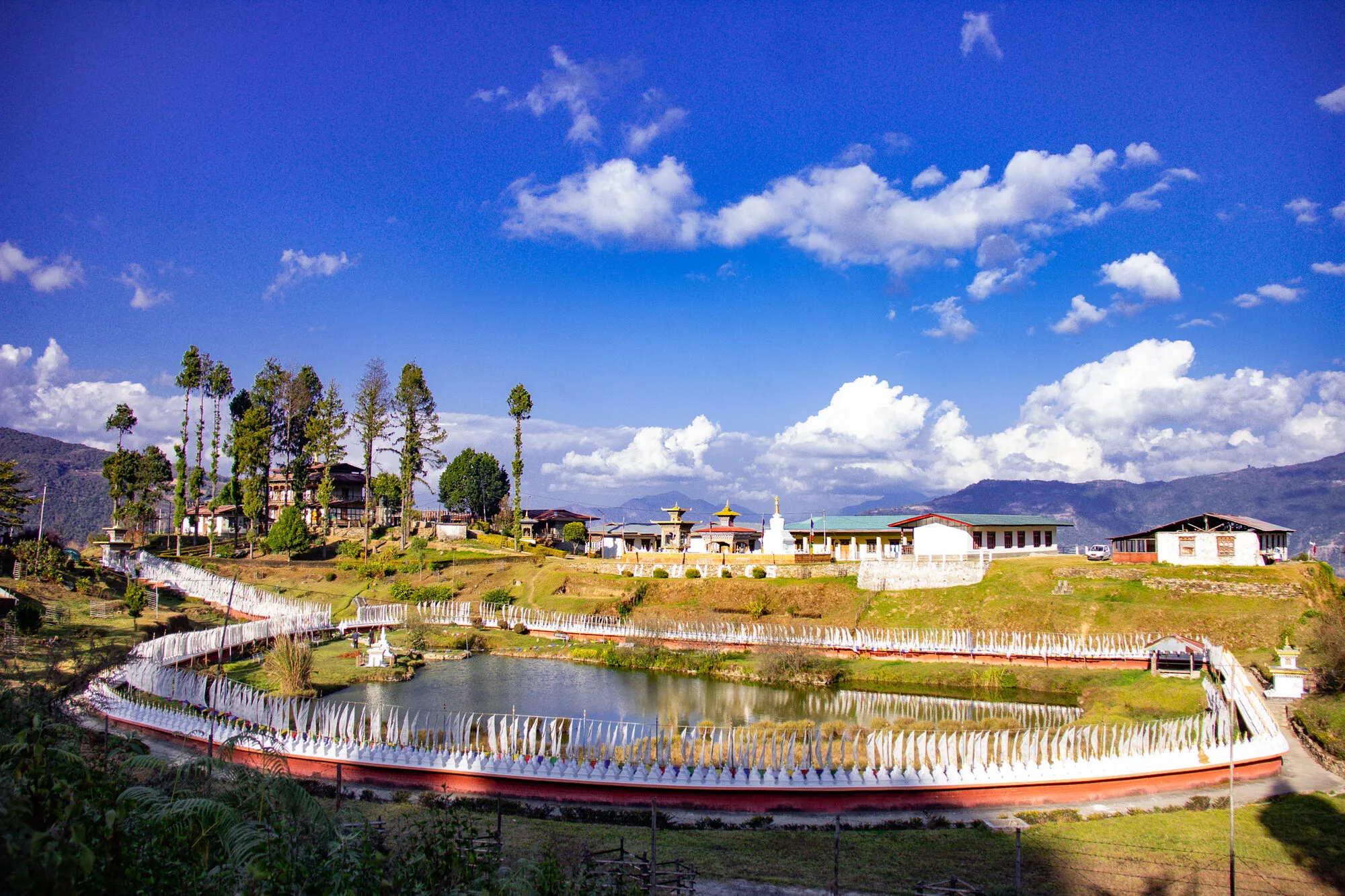 Yurung Tsho and its chorten wall below a terrace of school buildings and a small gold-roofed lhakhang, tall trees at the left